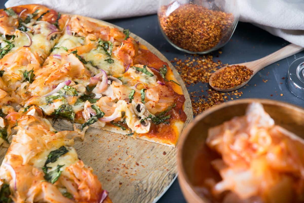 Sliced kimchi pizza on circular wooden cutting board, kimchi and chili pepper flakes on the side.