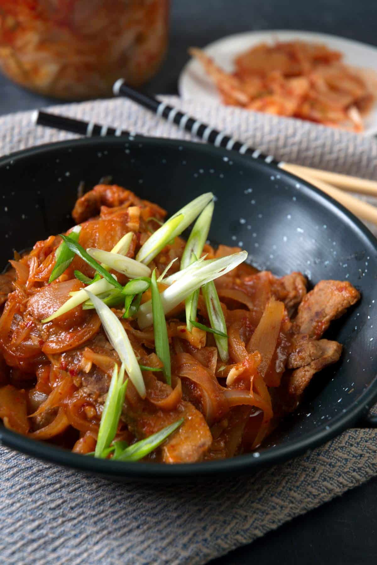 Kimchi stew in a black bowl with sliced scallions. Chopsticks and kimchi in the background.