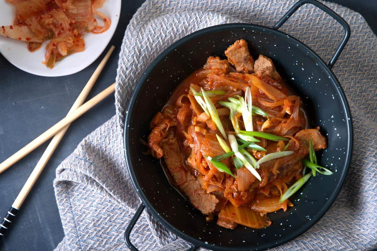 Kimchi stew in a black bowl with sliced scallions. Chopsticks and kimchi on the side.