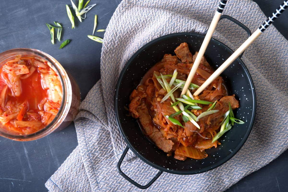 Kimchi stew in a black bowl with sliced scallions and chopsticks. Kimchi on the side.