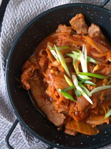 Kimchi stew in a black bowl with sliced scallions. Chopsticks and kimchi on the side.