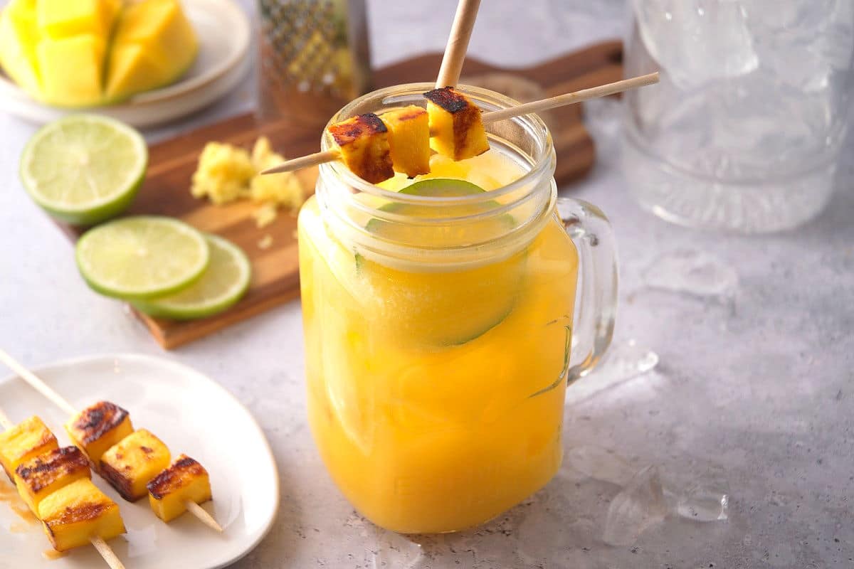 Mango cocktail with grilled mango skewer, straw and limes slices in a glass jar.