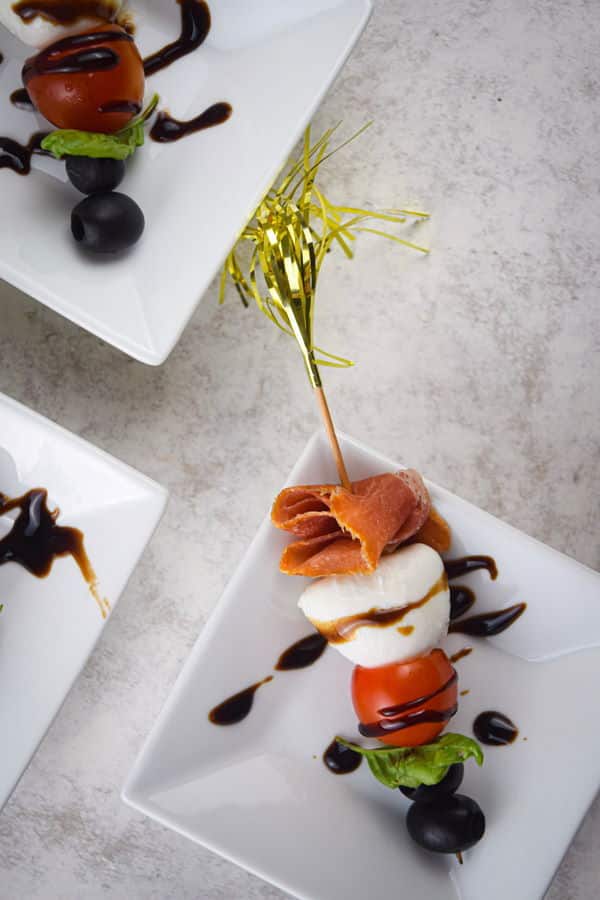 Caprese and prosciutto mini skewers on white dishes with balsamic drizzle.