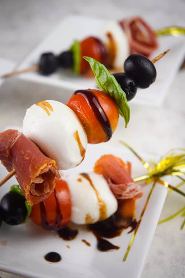 Caprese and prosciutto mini skewers on a white dish with balsamic drizzle.