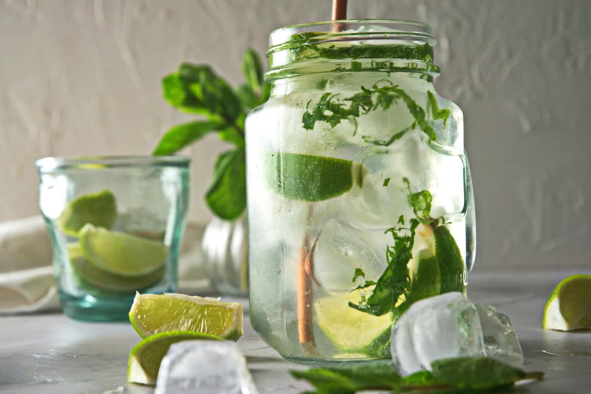 Front view of a mojito mocktail in a glass mug with a brown straw. Lime wedges and ice cubes on the side.