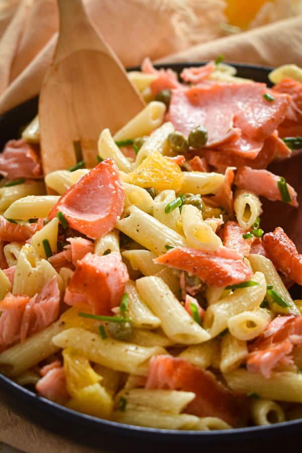 Smoked salmon pasta salad in a dark bowl with a wooden spoon and off-white dish cloth.