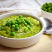 Pea purée in a white bowl on wooden plate, with a spoon on the side