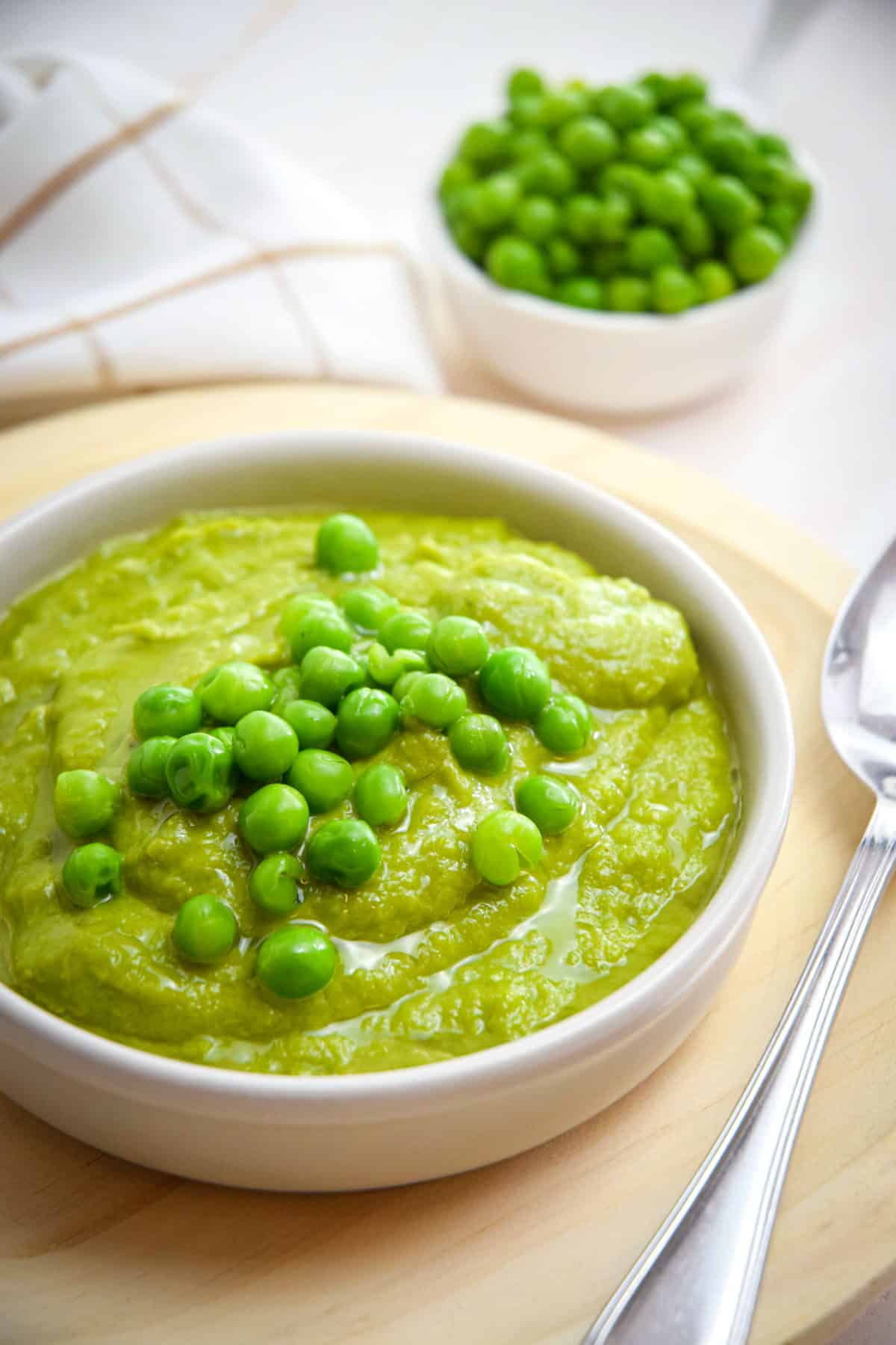 Pea purée in a white bowl on wooden plate, with a spoon on the side and green peas in the background.