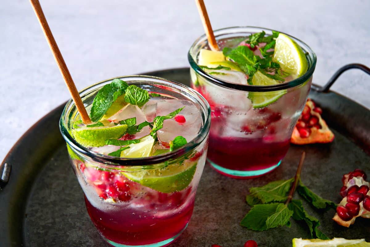 Pomegranate mojito mocktails on tray with mint leaves and pomegranate seeds on the side.