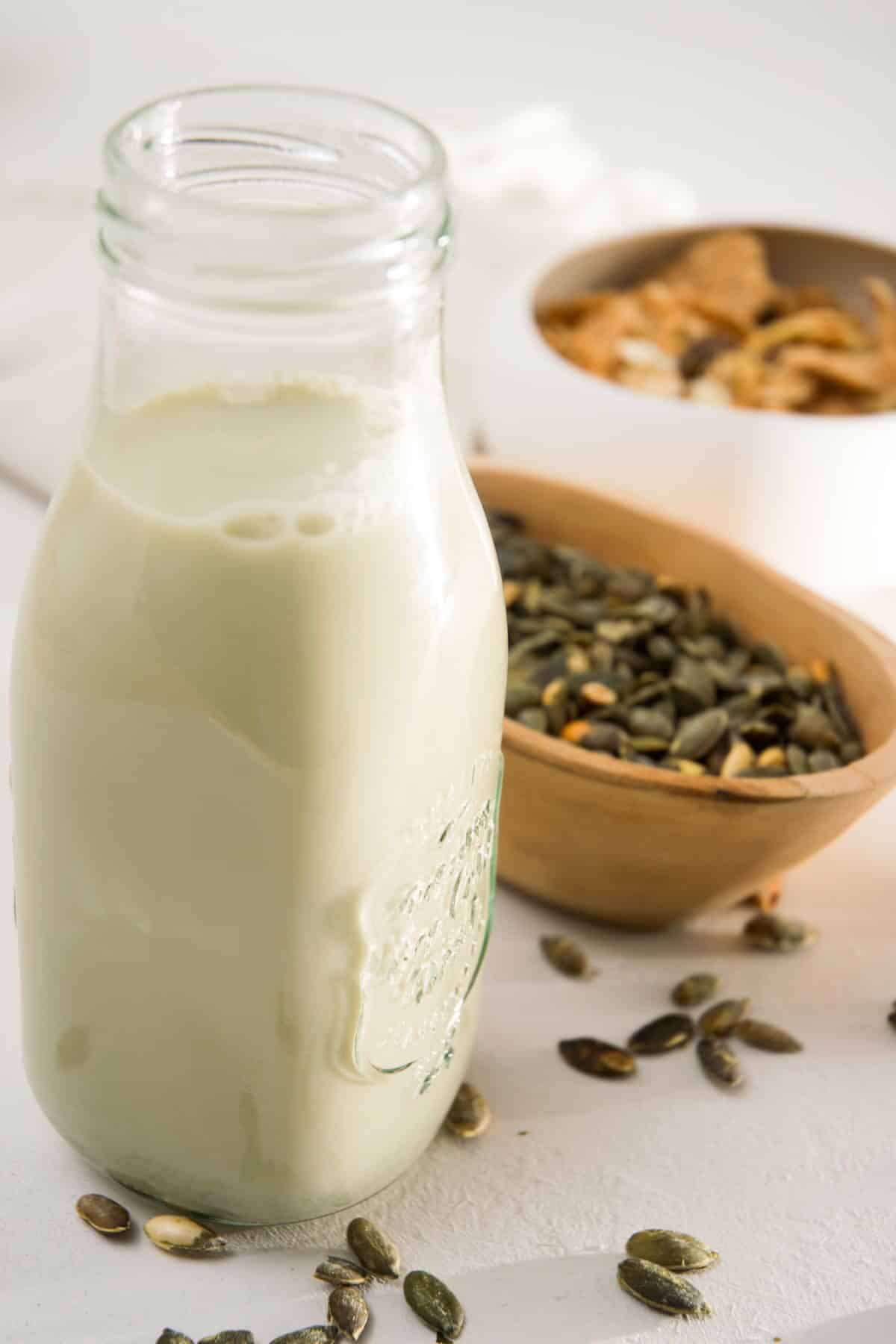 Pumpkin seed milk in a small jar on white background, a bowl of cereal and pumpkin seeds in the background.
