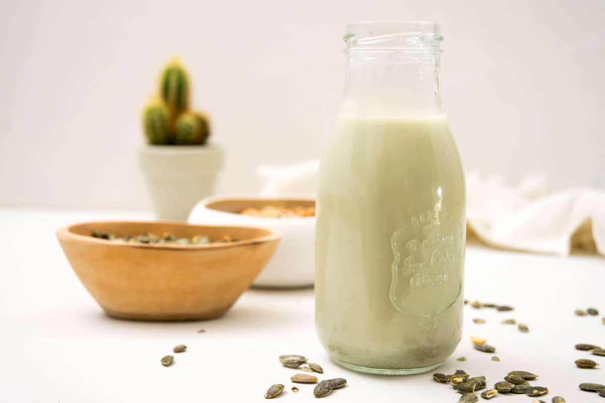 Pumpkin seed milk in a small jar on white background, a bowl of cereal and pumpkin seeds in the background.