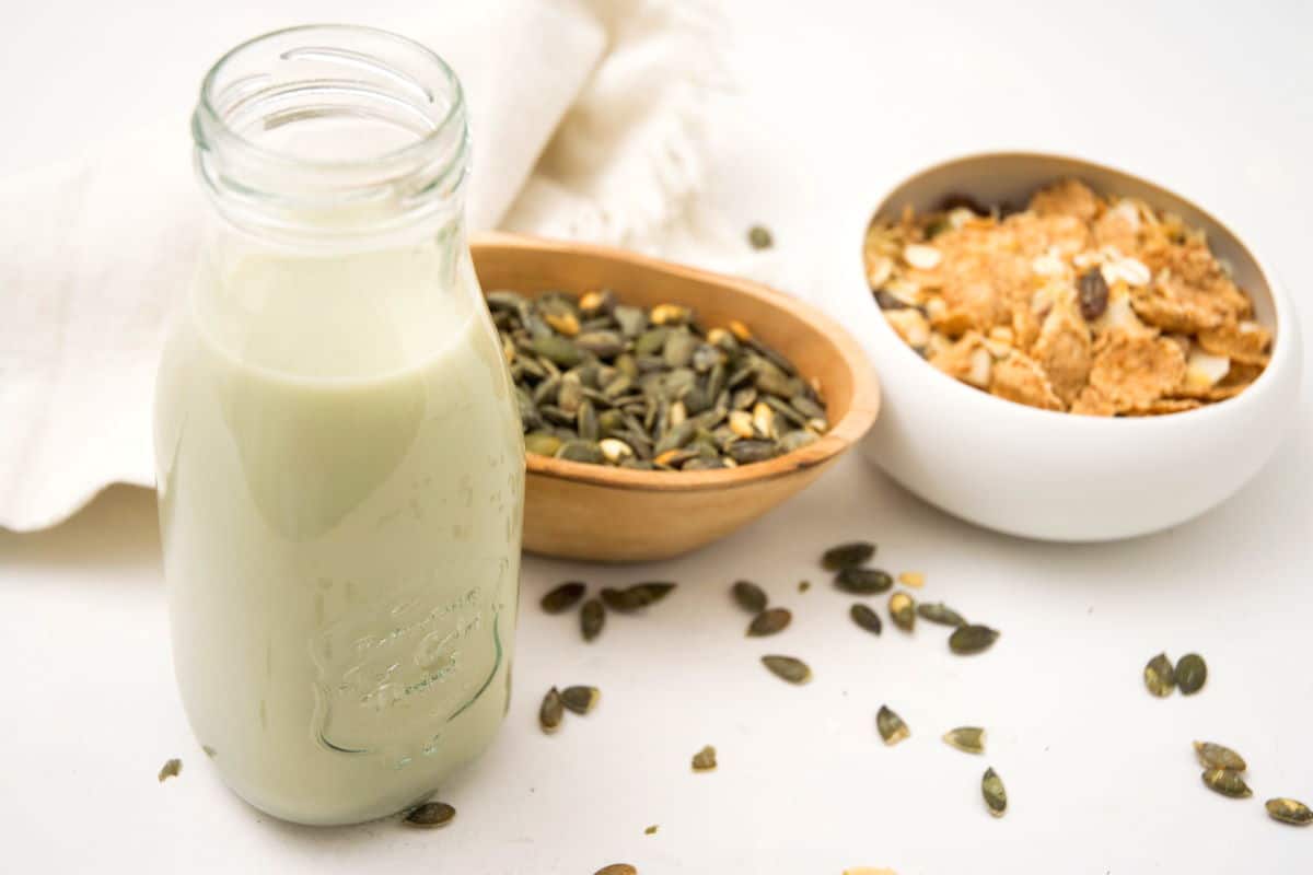Pumpkin seed milk in a small jar on white background, a bowl of cereal and pumpkin seeds in the background.
