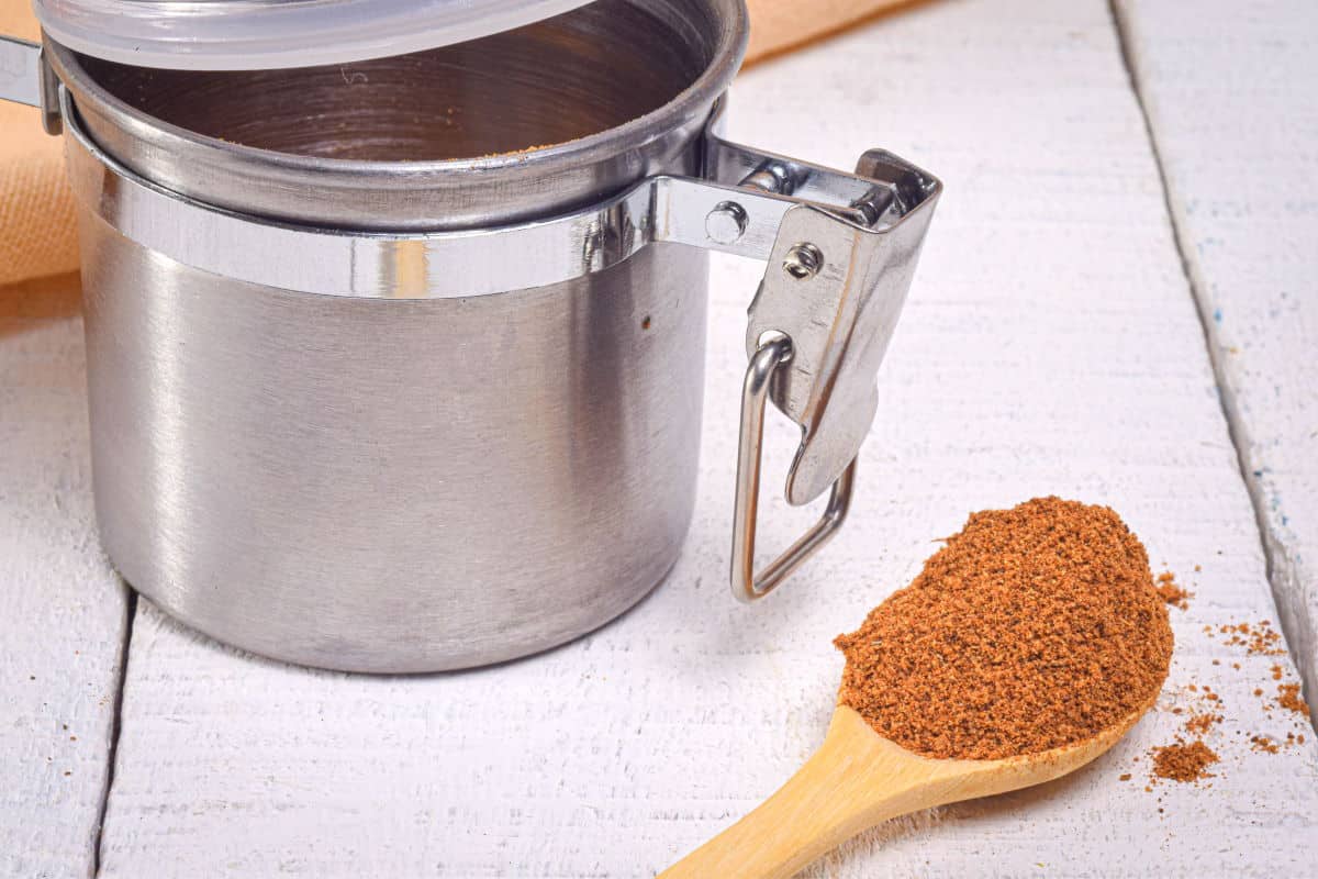 Pumpkin pie spice mix on wooden spoon beside metal spice container.