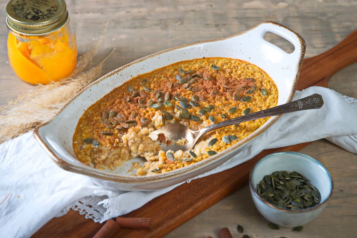 Pumpkin spice oatmeal in a white casserole dish with a spoon on wooden cutting board.