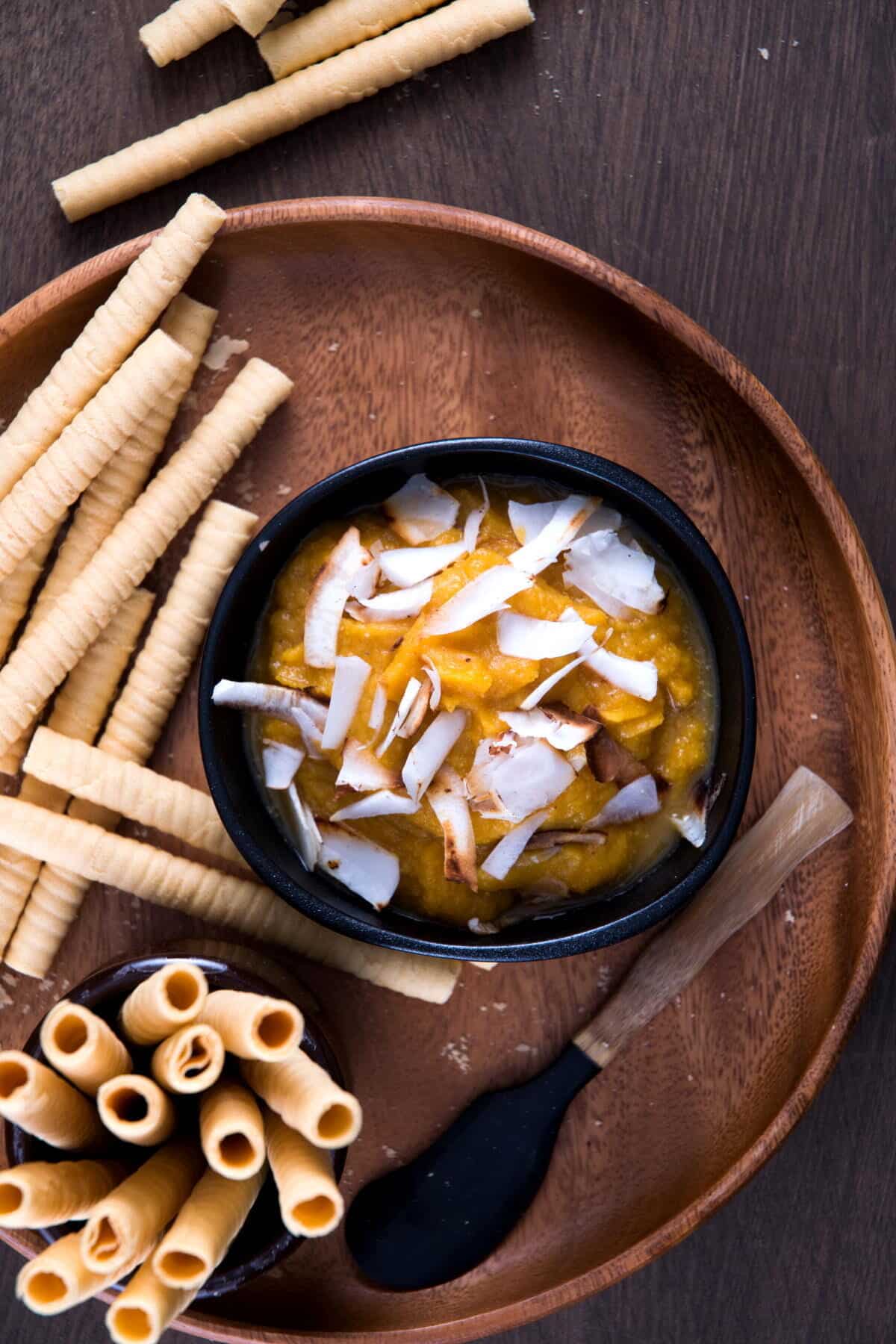 Pumpkin pie hummus in a black bowl on wooden plate with toasted coconut flake topping, breadsticks and small spatula on the side.