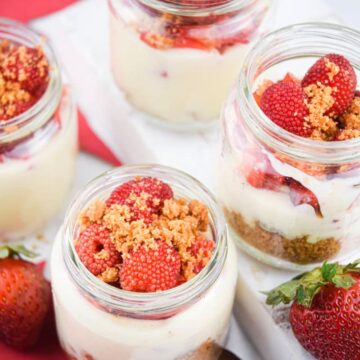 Raspberry cheesecake in mini dessert jars with strawberries surrounding the jars, white background.