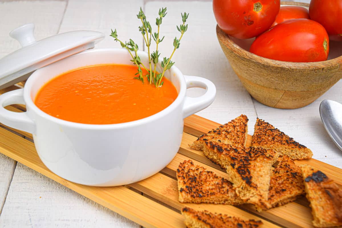 Roasted tomato soup in white bowl with toast points and Roma tomatoes on the side.