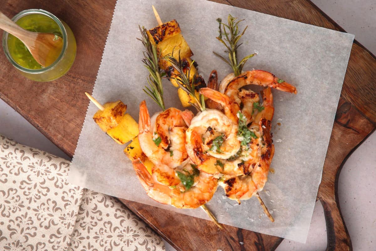 Top view of grilled shrimp on rosemary skewers and skewered pineapple wedges on parchment paper with sauce on the side.