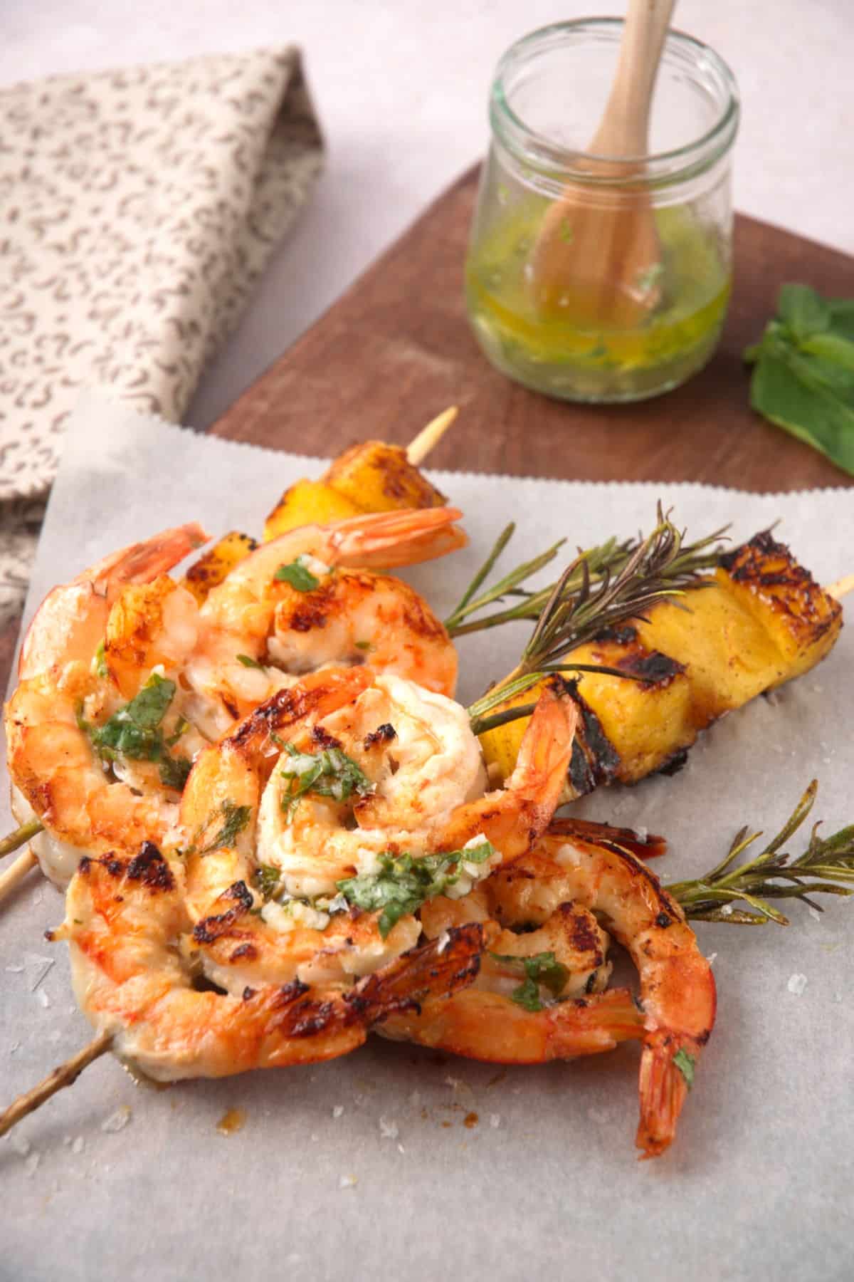Grilled shrimp on rosemary skewers and skewered pineapple wedges on parchment paper with sauce on the side.