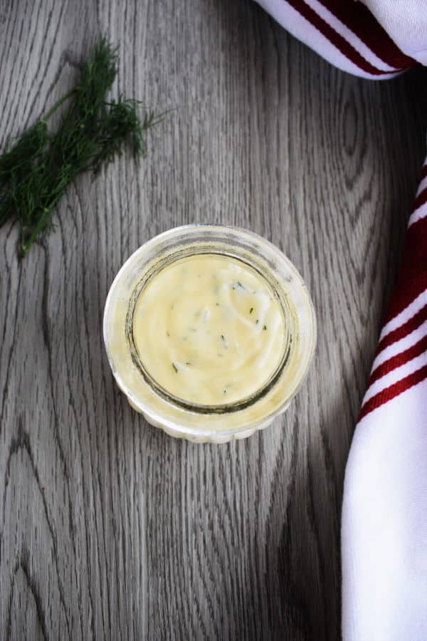 Honey dill sauce in a jar, kitchen towel and fresh dill on the side, grey wooden background.