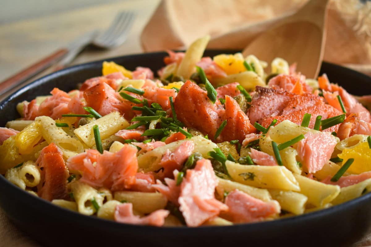 Smoked salmon pasta salad in a dark bowl with a wooden spoon.