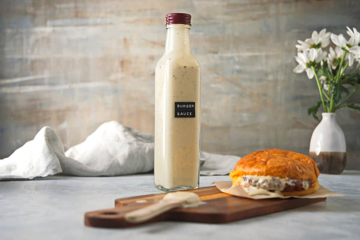 Burger sauce in a bottle with hamburger and spoon on cutting board. White flowers in the background.