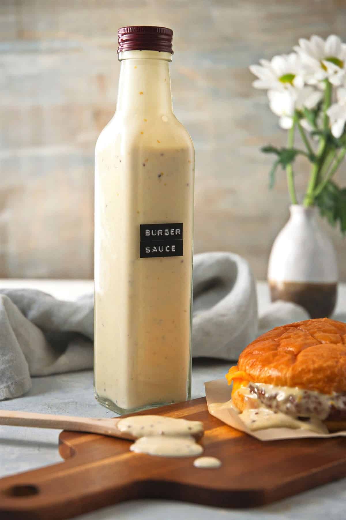 Burger sauce in a bottle with hamburger and spoon on cutting board. White flowers in the background.