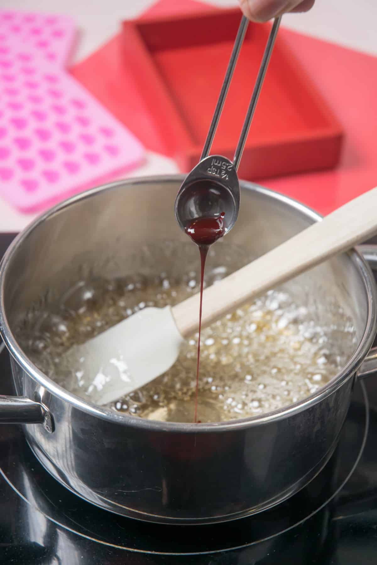 Candy mix in a saucepan with a spatula and liquid red food coloring.