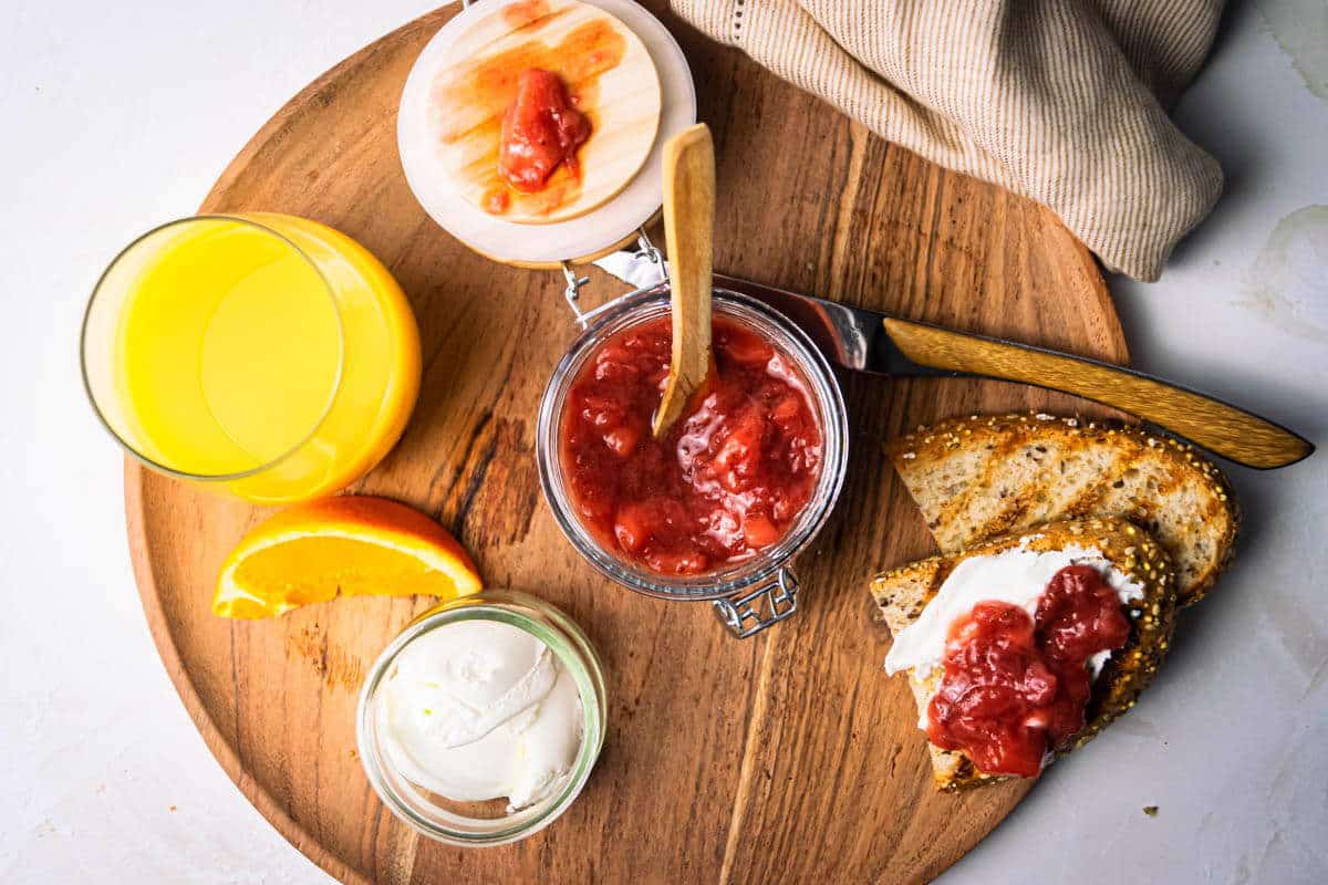 Strawberry compote in a jar, with toast and orange juice in a glass on the side, on wooden circular serving dish.