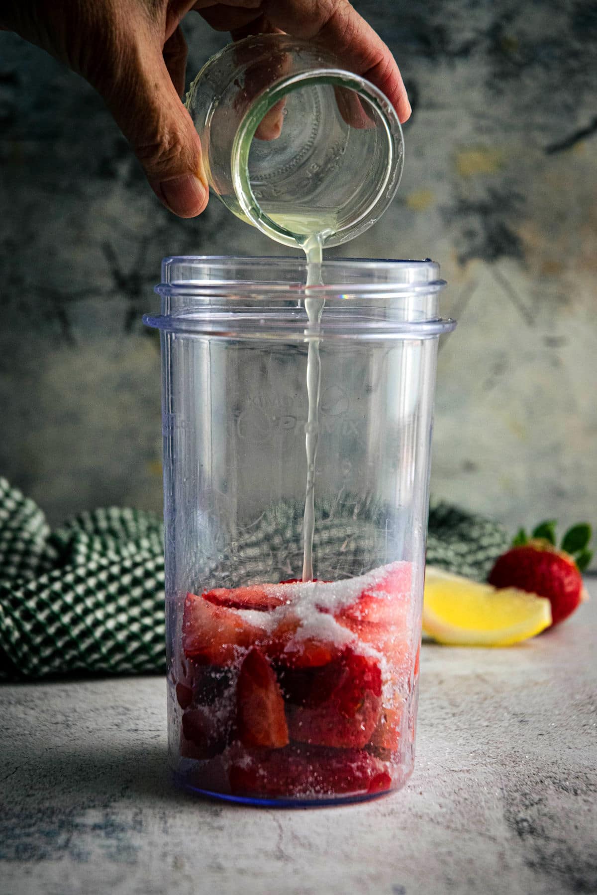 Strawberries and sugar in a blender jar. Lemon juice pouring into the jar.