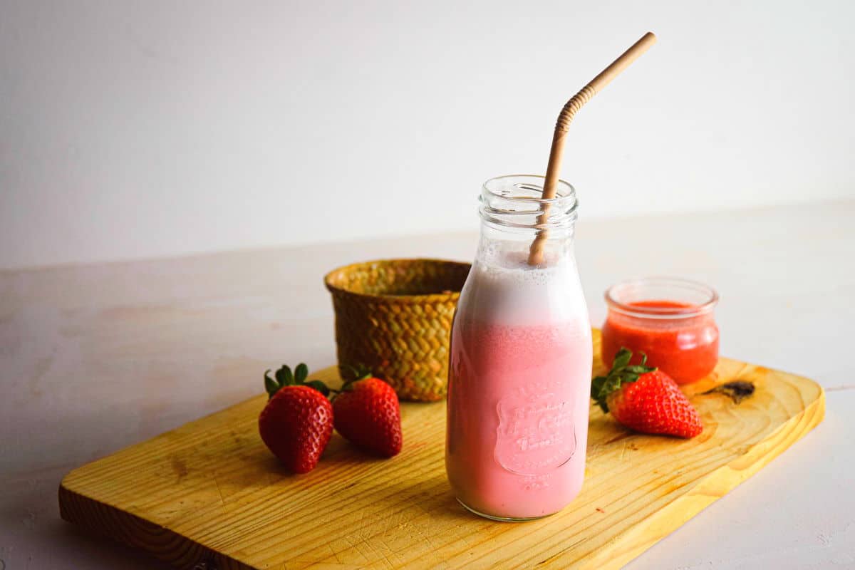 Strawberry Milk in a glass jar with a straw on a wooden board with fresh strawberries and strawberry puree.
