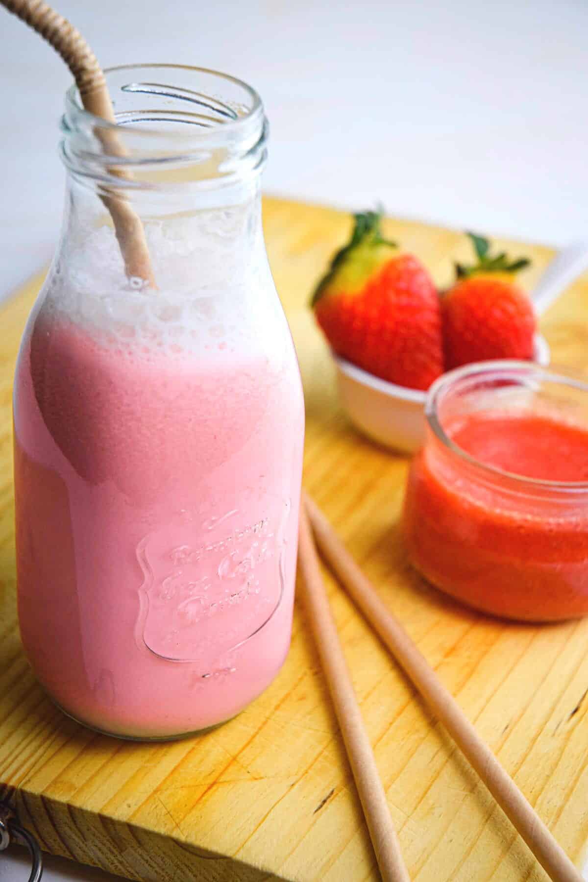 Strawberry Milk in a glass jar with a straw on a wooden board with fresh strawberries and strawberry puree.