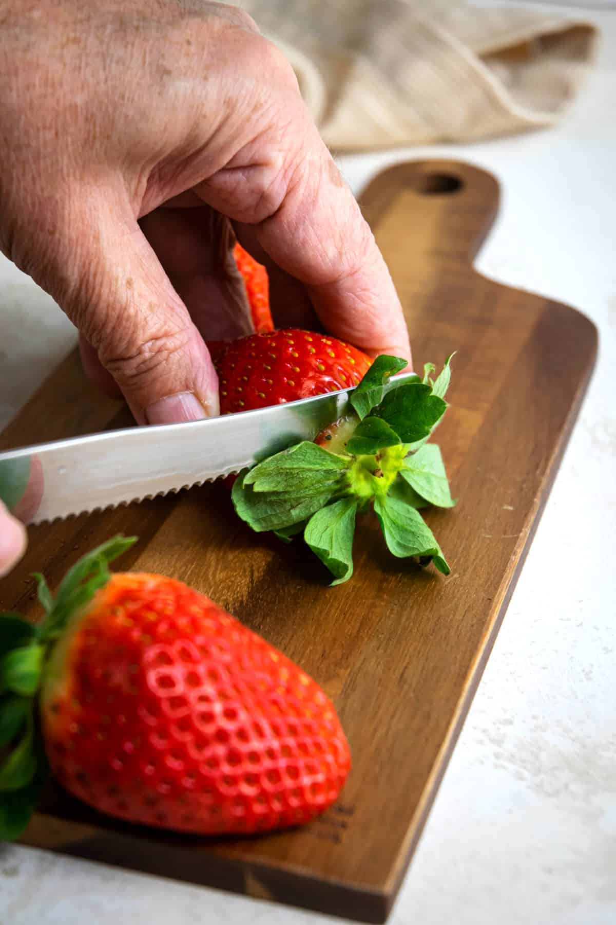 Strawberries getting tops cut off on wooden cutting board.