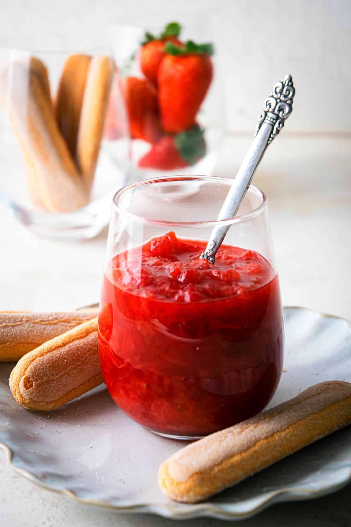Strawberry-rhubarb sauce in a glass with a spoon and biscuits on the side.