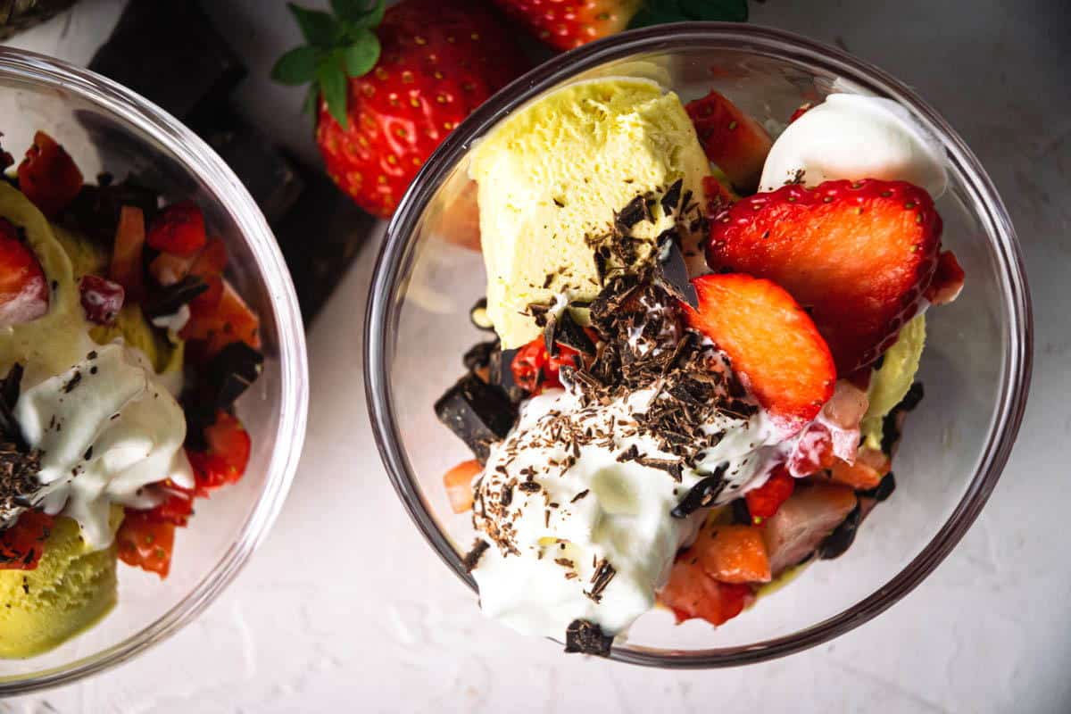 2 strawberry sundaes in dessert bowls, chocolate pieces and strawberries on the side.