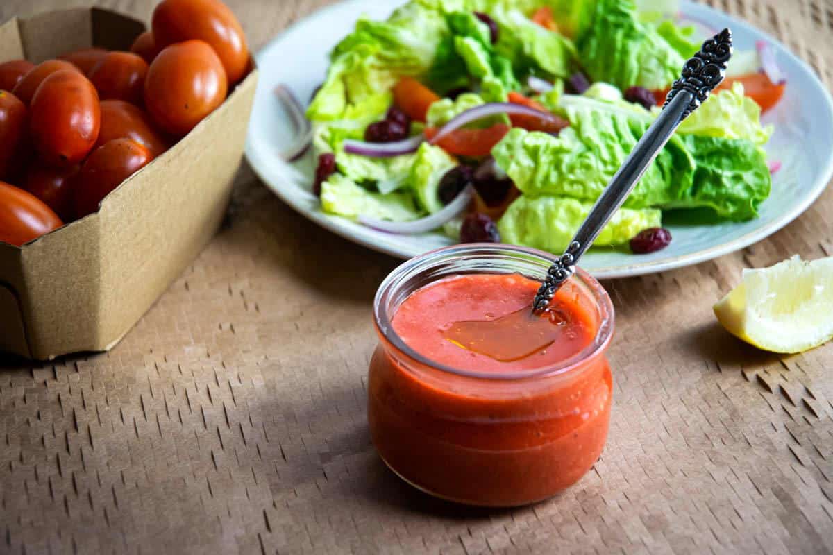 Strawberry vinaigrette in a jar with a spoon, fresh salad and tomatoes in the background.