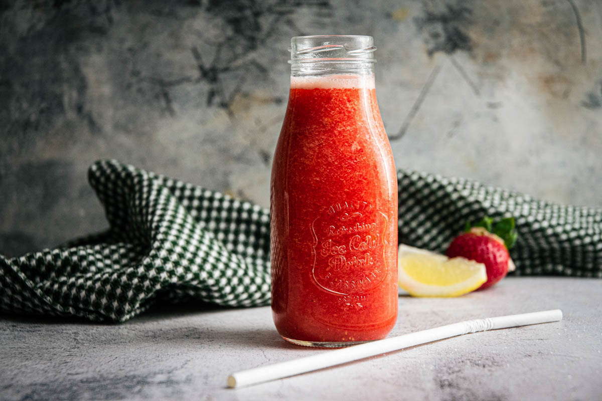 Strawberry juice in a glass jar, with a straw, lemon wedge and strawberry on the side.