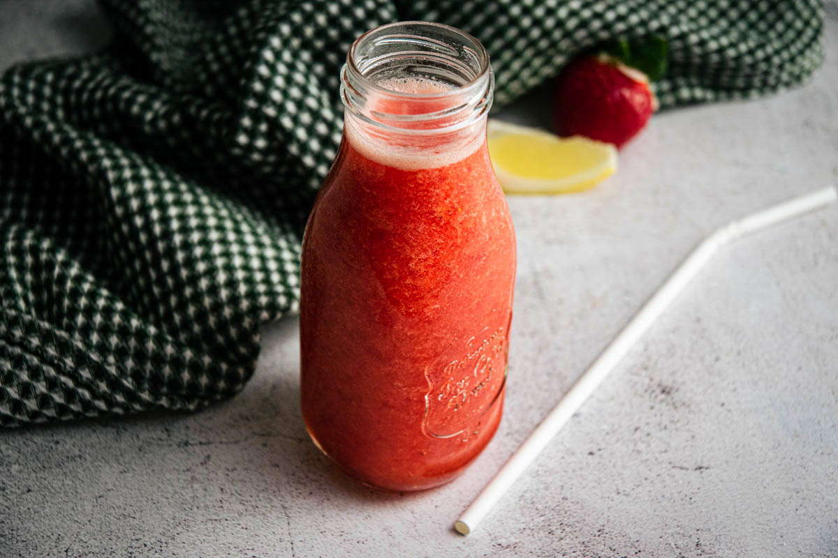 Strawberry juice in a glass jar, with a straw, lemon wedge and strawberry on the side.