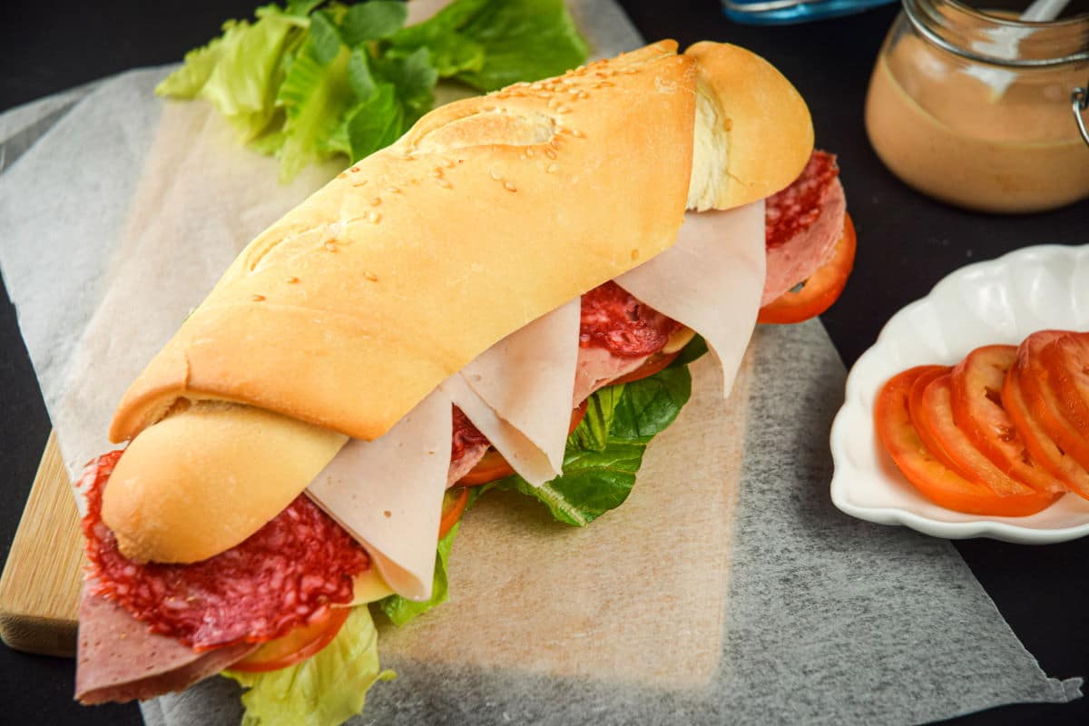 Triple meat sub sandwich with sriracha sauce on wooden cutting board and deli paper.