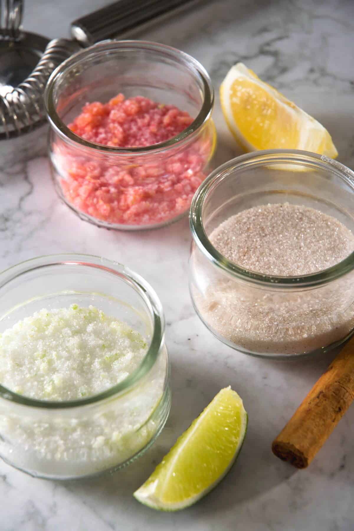Sugar rims in 3 small jars, a lime wedge, lemon wedge and cinnamon stick on the side.