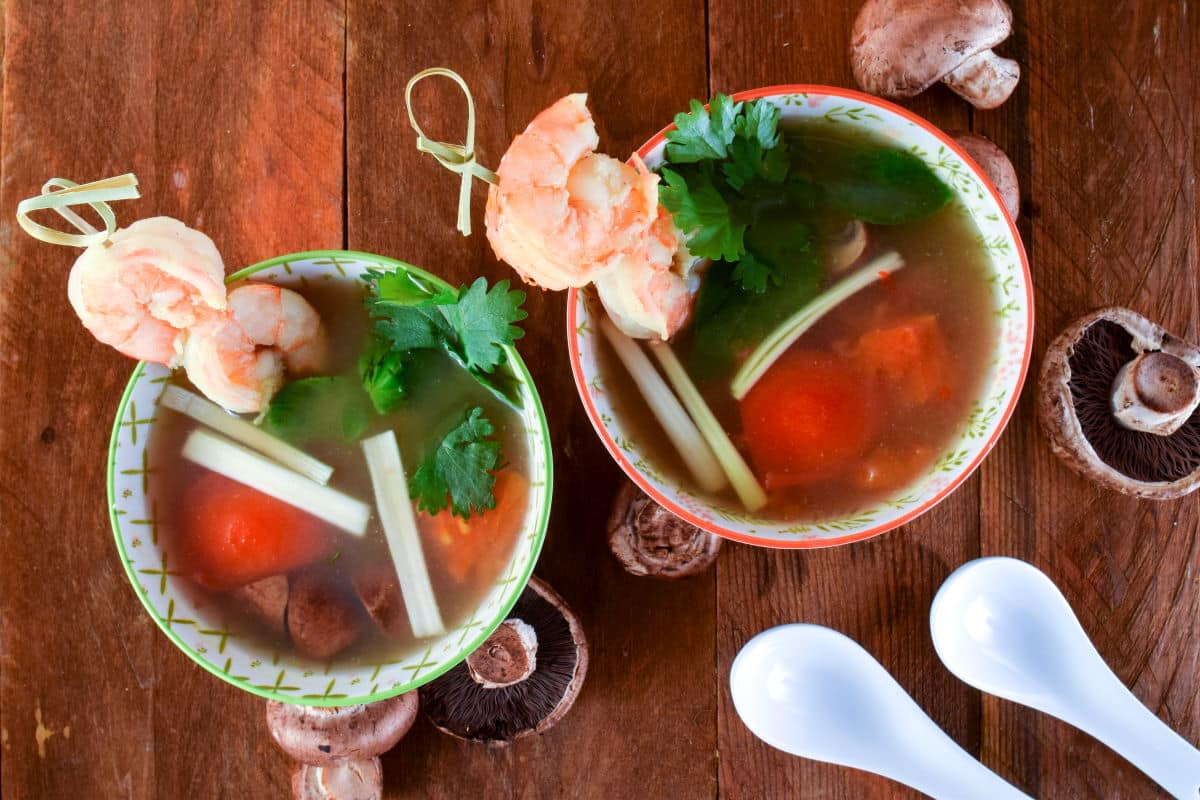 Top view of Tom Yum Soup in porcelain bowls with mushrooms and soup spoons on the side.