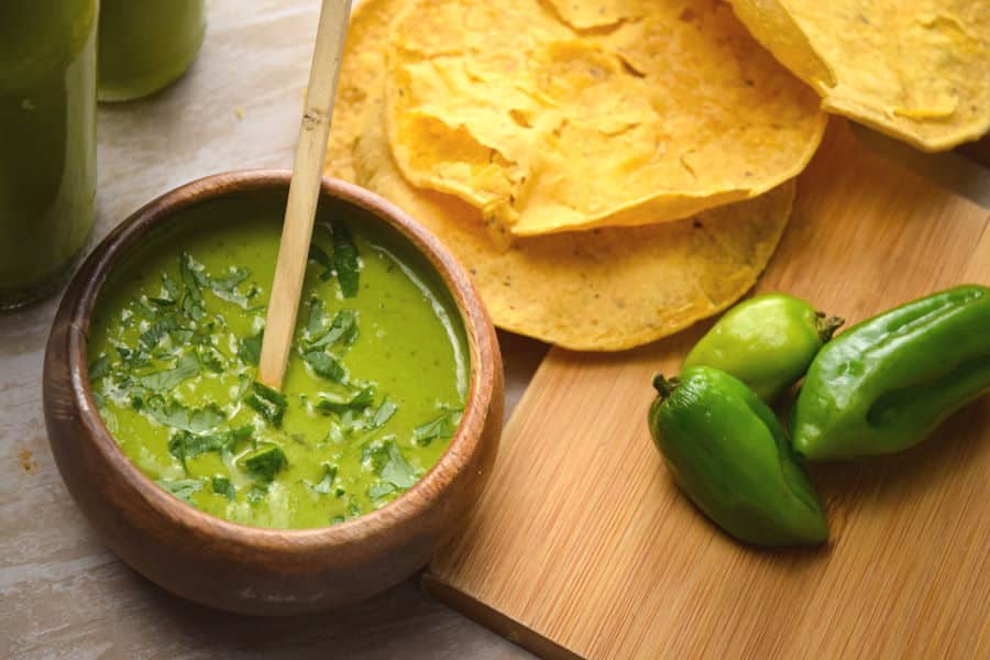 Tomatillo hot sauce in a wooden bowl, tortillas and peppers on the side.