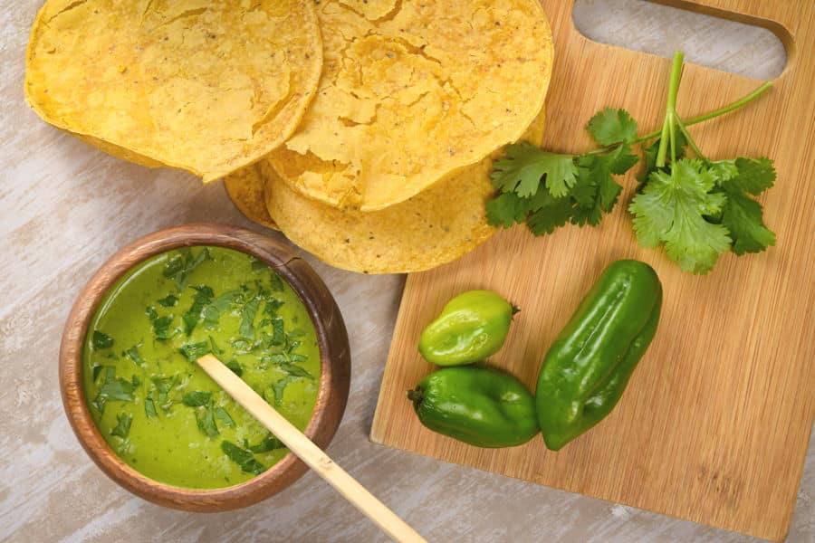 Tomatillo hot sauce in a wooden bowl, tortillas, cilantro and peppers on the side.