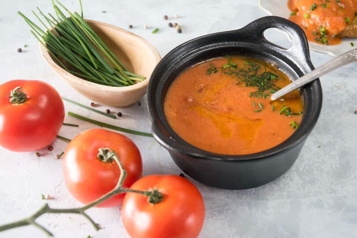 Tomato gravy in a small cast iron bowl, tomatoes on the vine and chives on the side.