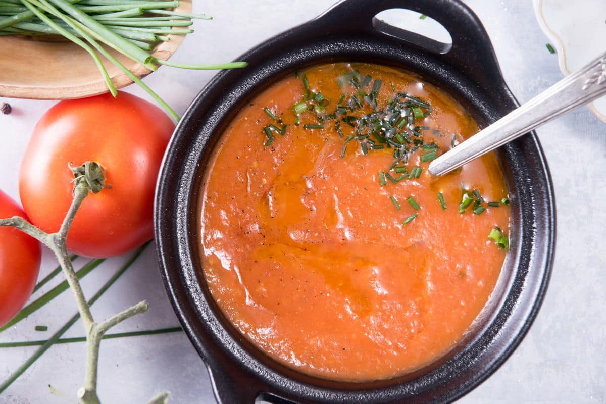 Tomato gravy in a small cast iron bowl, tomatoes on the vine and chives on the side.