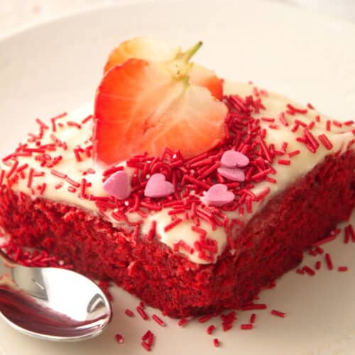 A piece of red velvet sheet cake with red and pink candy decorations and sliced strawberries on white plate.