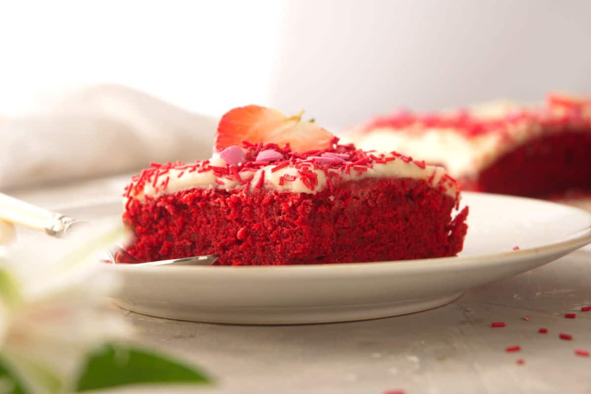 A piece of red velvet sheet cake with red and pink candy decorations on white plate.