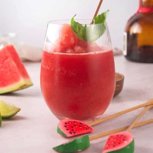 Watermelon basil cocktail in wine glass with watermelon stir sticks and lime wedges on the side.