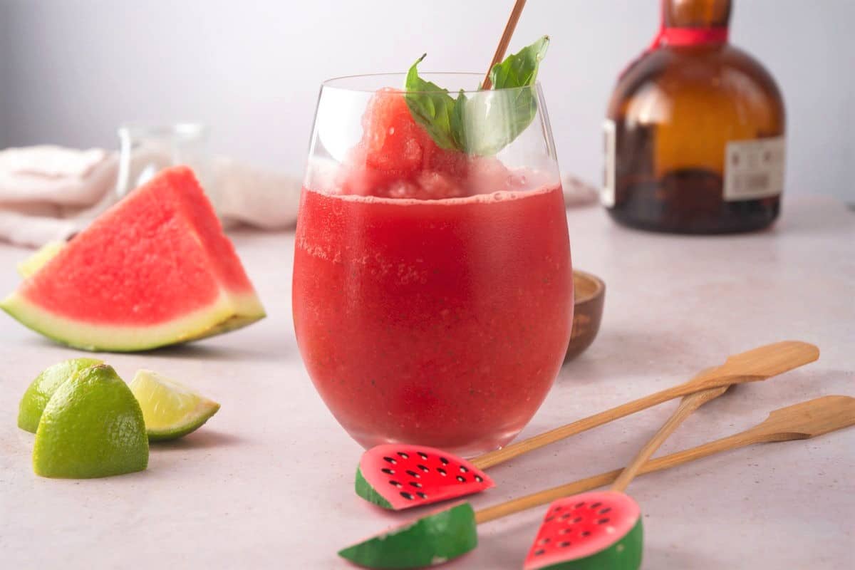 Watermelon basil cocktail in wine glass with watermelon stir sticks and lime wedges on the side.
