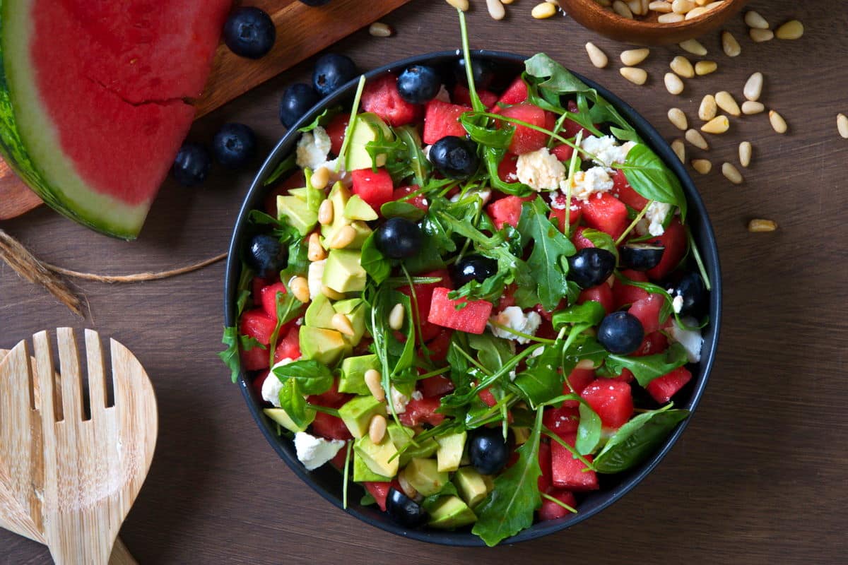 Top view of watermelon salad in a black bowl on wooden background. Salad spoons, pine nuts, blueberries and watermelon on the side.